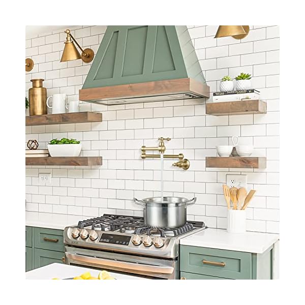 This image showcases a stylish kitchen setup with a brass pot filler faucet installed above a stove, filling a pot directly. The kitchen design features a farmhouse aesthetic, with green cabinets, wooden open shelves, and white subway tile backsplash. The brass elements, including the faucet and wall-mounted lights, add a touch of elegance and warmth, complementing the green and wood tones throughout